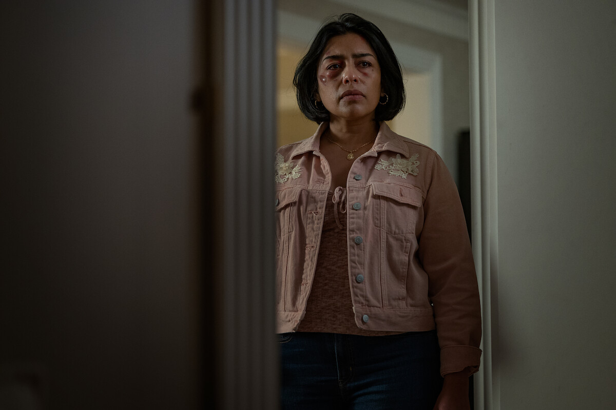 Woman with bruised eyes stands indoors near a doorway, wearing a pink jacket and looking seriously ahead, in a dimly lit home environment.