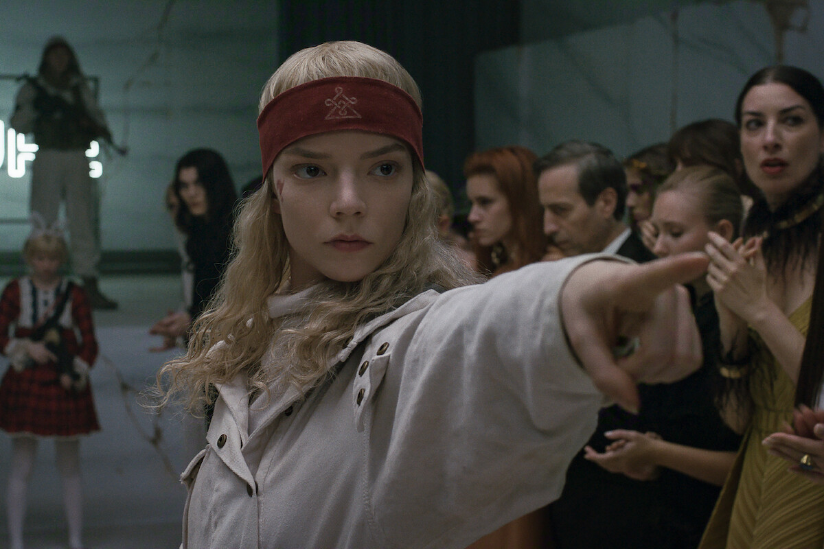 A young woman with blonde hair and a red headband points assertively in a tense, modern indoor setting. Several people dressed in eclectic, stylish clothing watch her intently, suggesting a dramatic or intense situation.