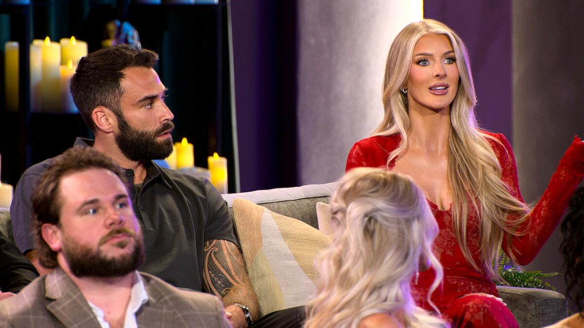 Four people are sitting on couches indoors, dressed formally. Candles in the background create a warm, intimate setting. One woman in a red dress is speaking while others listen, suggesting a discussion or interview atmosphere.