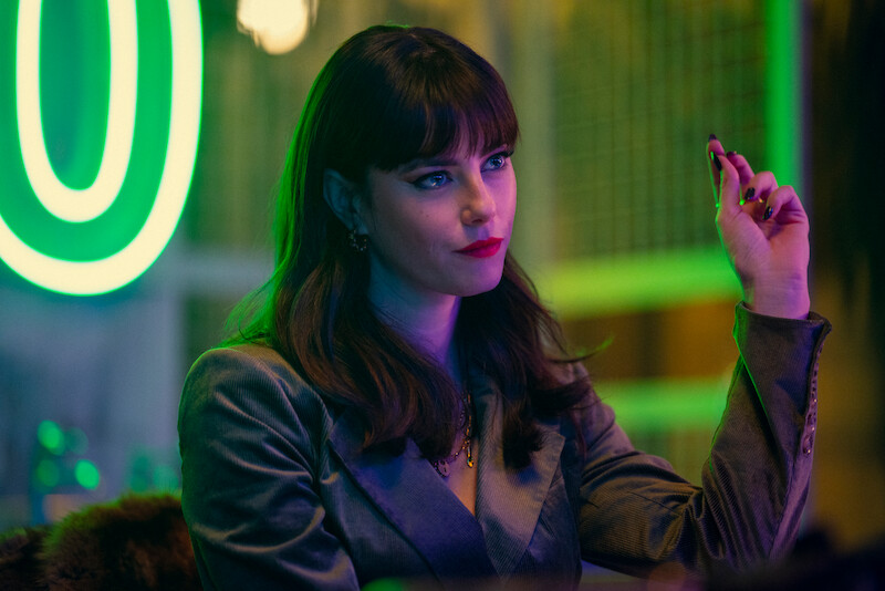 Kaya Scodelario wears a gray jacket and is shrouded in green neon light in Season 1 of ‘The Gentlemen.’
