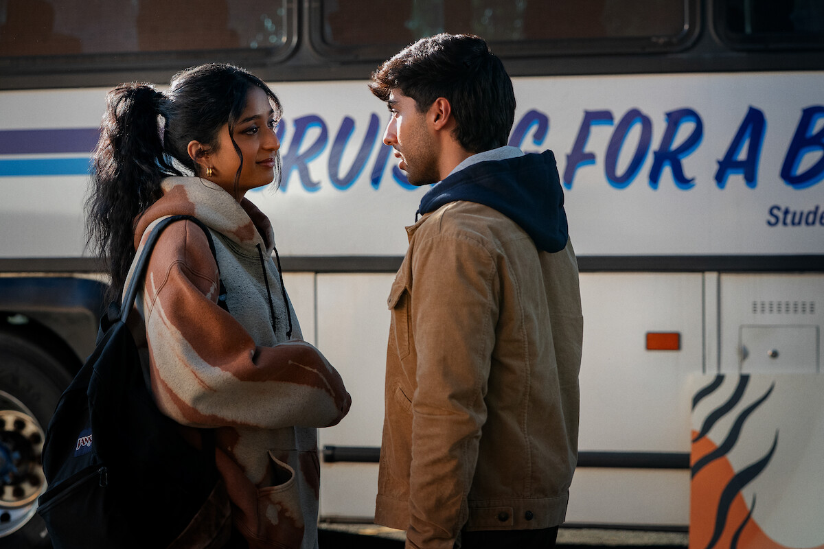 Two young adults stand facing each other at a bus stop, engaged in a serious conversation. There is a large bus with partial text in the background, suggesting a travel or departure scene.