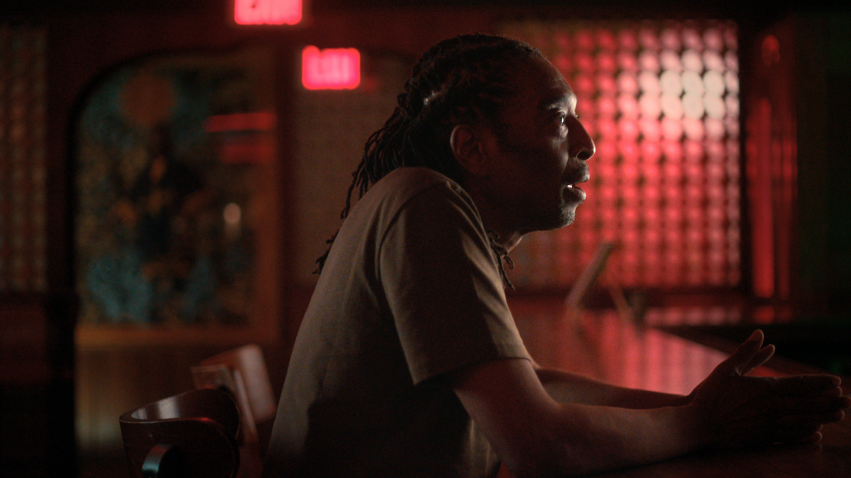 Man with dreadlocks sits at a dimly lit bar, illuminated by red light. The background features EXIT signs and warm decor, creating a moody, contemplative atmosphere.