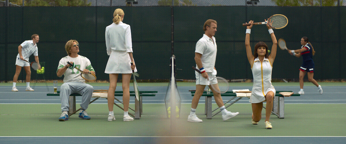 People in white tennis outfits on a court; some are seated or standing by benches, one woman lunges with a racket raised, others play in the background on a bright outdoor court with a green fence.