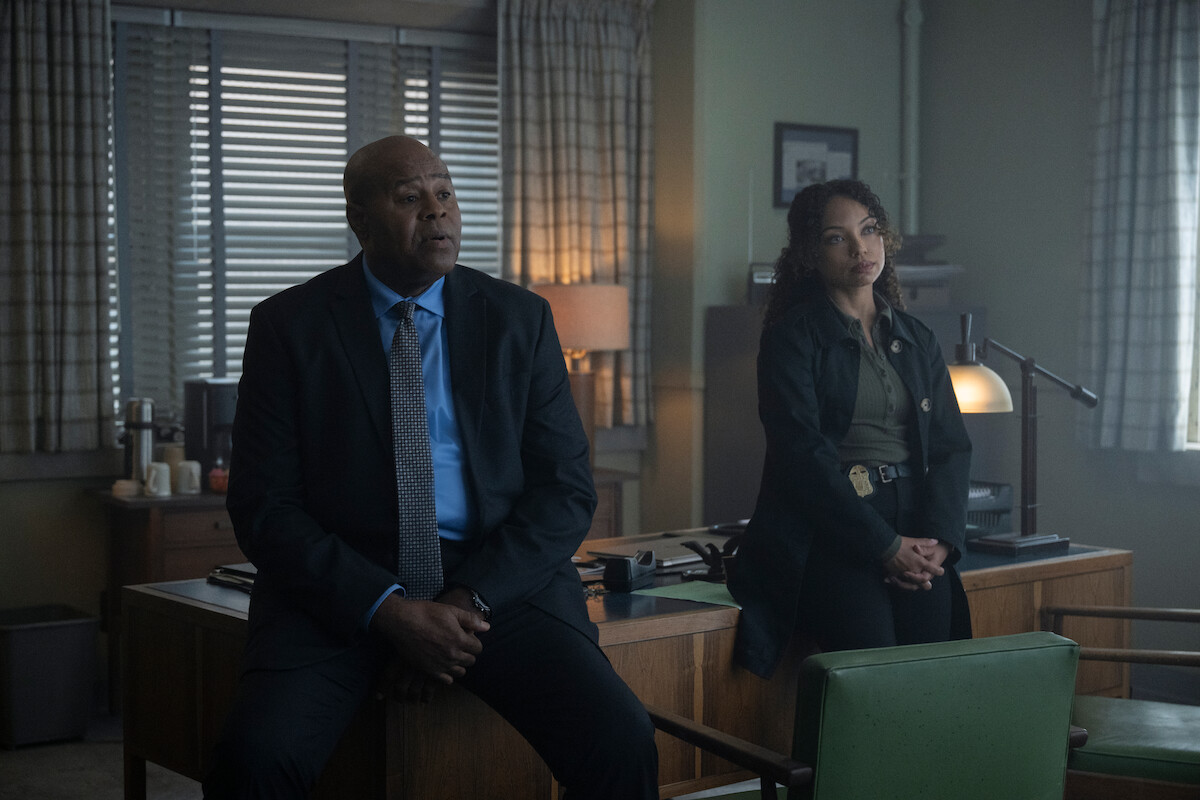Two detectives sit and lean against a wooden desk in a dimly lit office with large windows, blinds, lamps, and paperwork, suggesting a serious conversation or investigation.