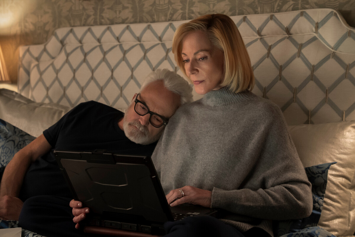 Older couple relaxing in bed together, man resting his head on woman's shoulder while she uses a tablet, cozy bedroom setting with patterned headboard and soft lighting.
