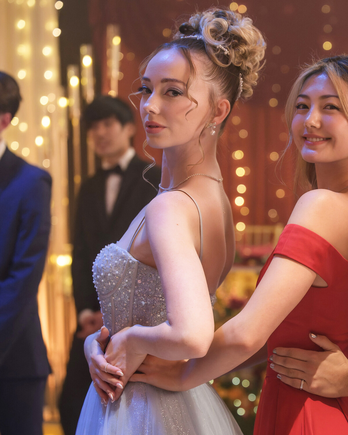 Two women in elegant dresses pose together at a formal event, surrounded by festive lights and decor. The focus is on their smiling faces and embrace, with other guests in formal attire visible in the softly lit background.