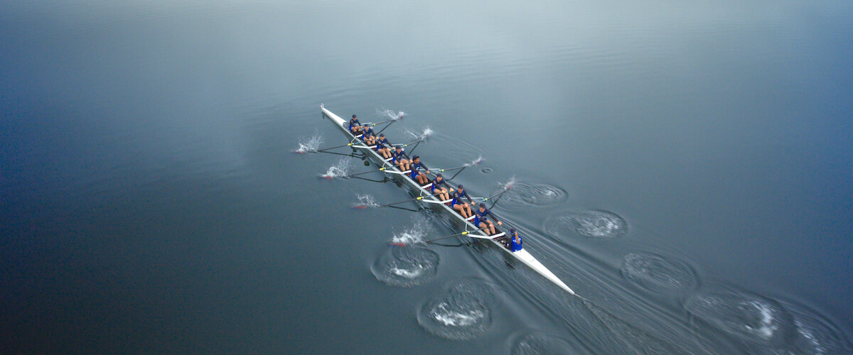 Aerial view of a rowing team in a long boat moving in sync across calm, reflective water, creating ripples as they row together in a misty, tranquil outdoor environment.
