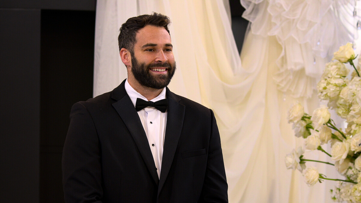 Man in a black tuxedo smiling at a formal indoor event, likely a wedding, with white flowers and draped white fabric in the background.