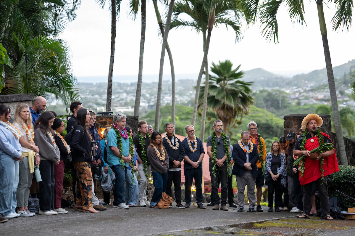 A group of people stands outdoors in a tropical setting with palm trees, listening to a person in traditional clothing holding a conch shell, with mountains and cityscape in the background.