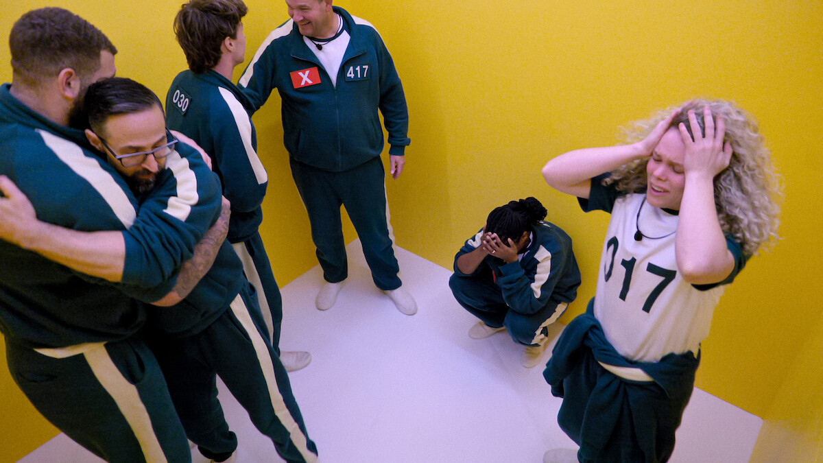 Six people in numbered tracksuits stand in a small yellow room; some appear distressed or emotional, with two hugging and others holding their heads or crouching, suggesting tension or anxiety in a confined environment.