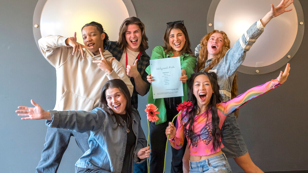 Group of six happy young adults posing energetically indoors in front of a grey wall with circular lights, one person holding up a certificate and others smiling and gesturing enthusiastically.