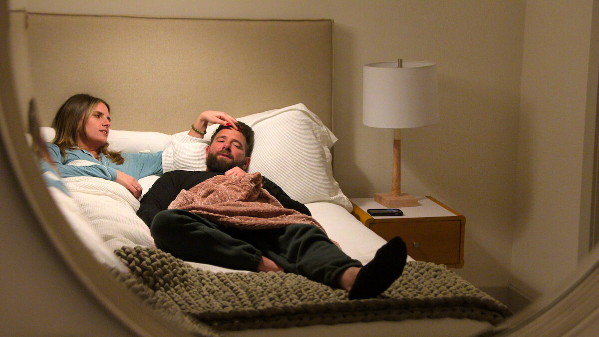Two people relaxing on a bed in a cozy, softly lit bedroom with beige walls, a lamp on a nightstand, and a blanket, seen through a round mirror.