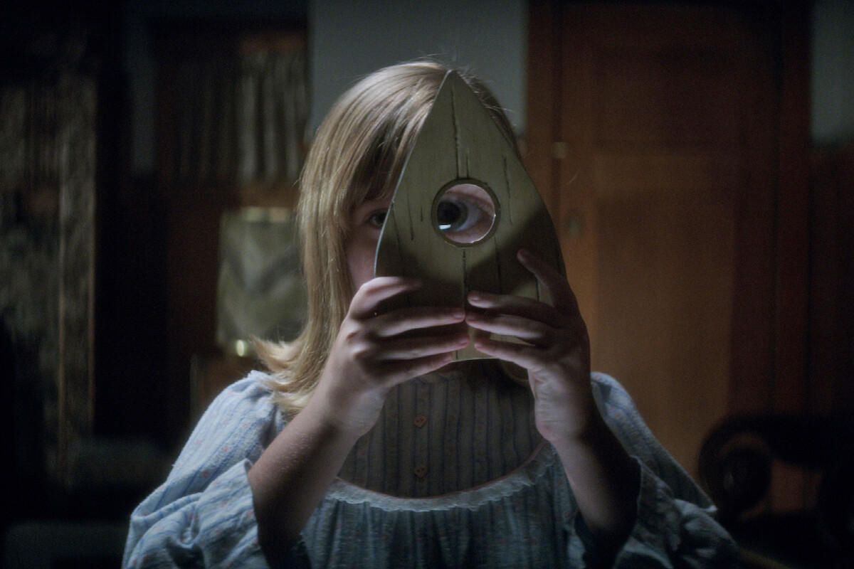 A young girl in a nightgown holds a wooden Ouija board planchette up to her eye in a dimly lit, old-fashioned room with wooden furniture and soft lighting.