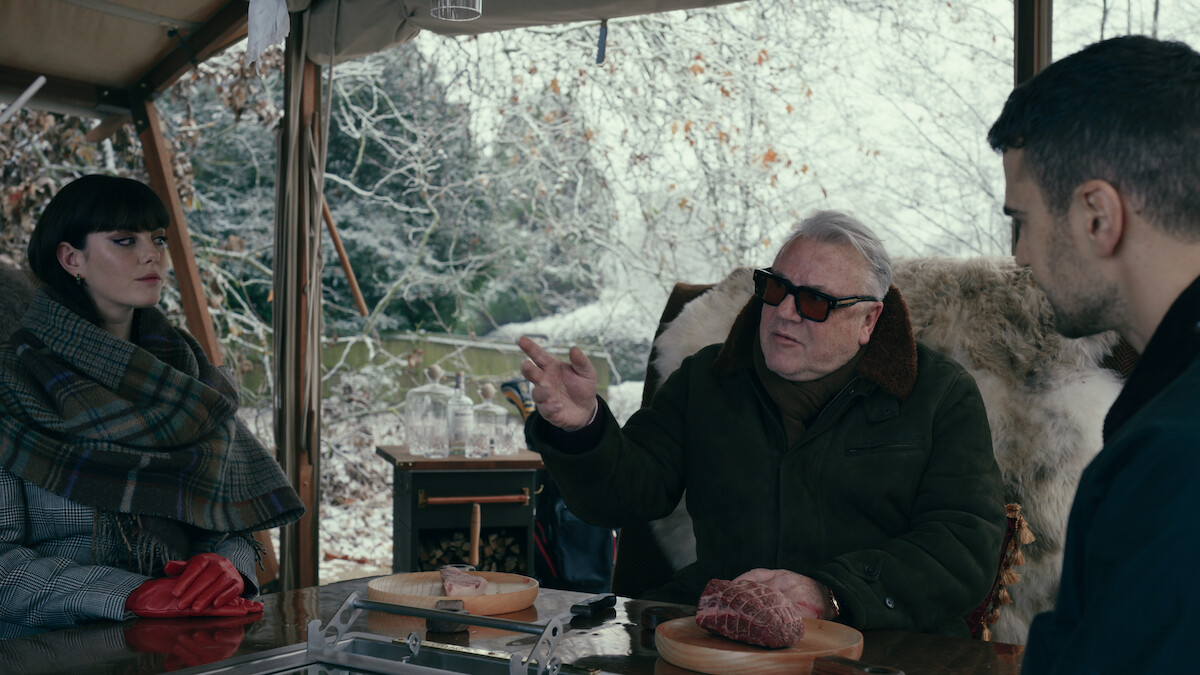 Kaya Scodelario as Susie Glass, Ray Winstone as Bobby Glass, and Theo James as Eddie Horniman dine at a table outdoors together in Season 1 of ‘The Gentlemen’