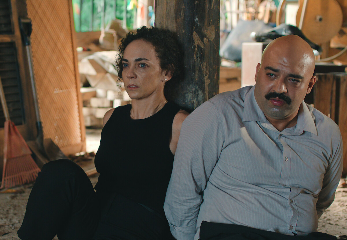 Duas pessoas, uma mulher branca de cabelos encaracolados e um homem branco, careca e de bigode, estão sentados no chão e amarrados a uma pilastra