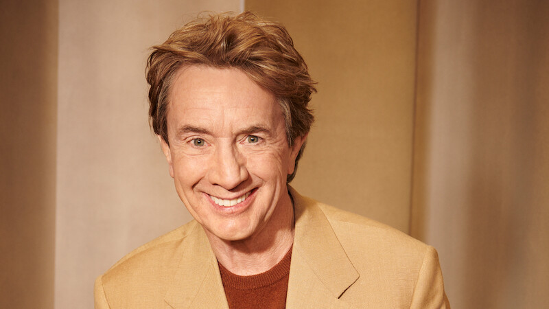 Martin Short smiles at the camera wearing a tan jacket.