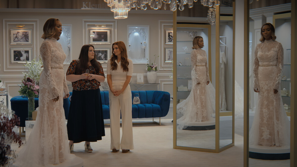 Three women in a stylish bridal shop, one wearing a wedding gown standing by mirrors, with elegant decor, framed photos on the wall, a blue couch, and chandeliers creating a classy atmosphere.