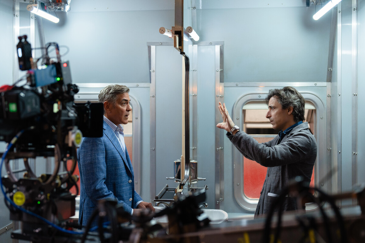 Two men converse seriously in a brightly lit, modern industrial room, with filmmaking equipment visible, suggesting a movie set. The mood is focused and professional.