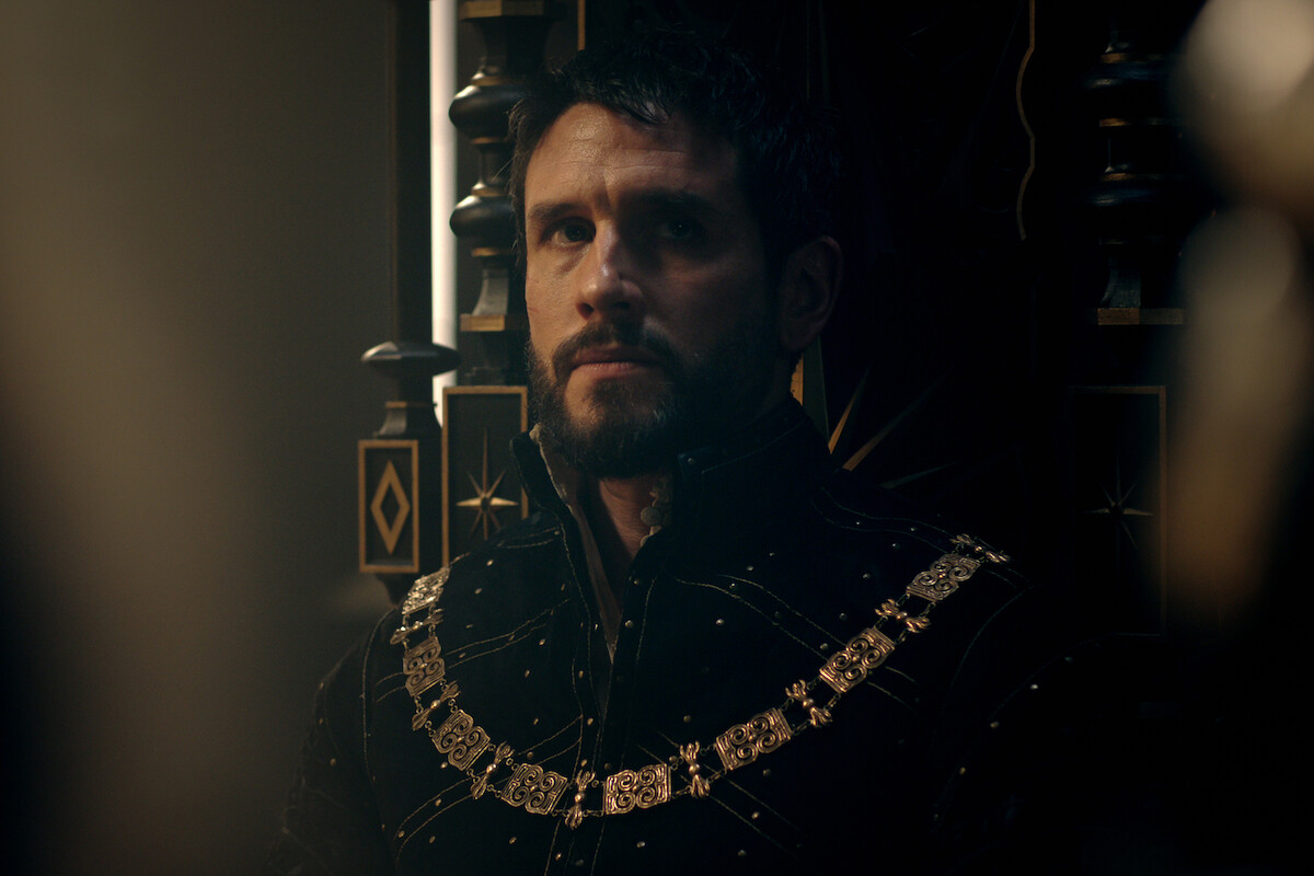 A stern man in ornate medieval clothing sits on a grand, dark throne in dim light, suggesting royalty or nobility in a regal, serious setting with a somber atmosphere.