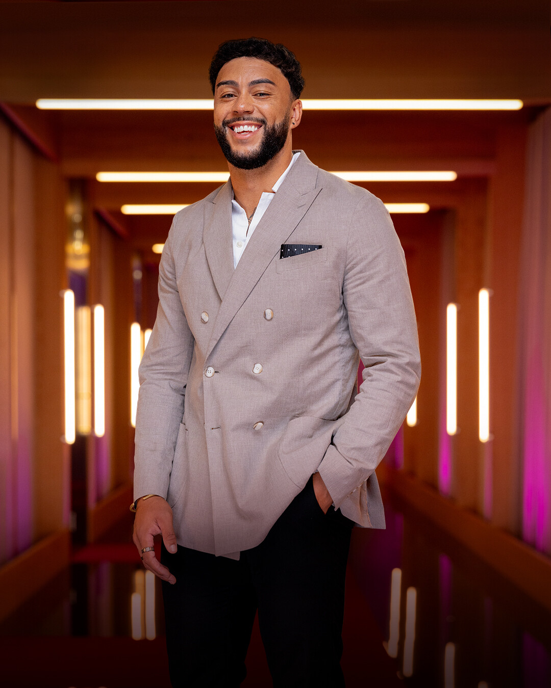 Man in a light beige suit jacket and black pants standing in a stylish, warmly lit hallway with glowing vertical lights and a modern interior design, smiling with one hand in his pocket.