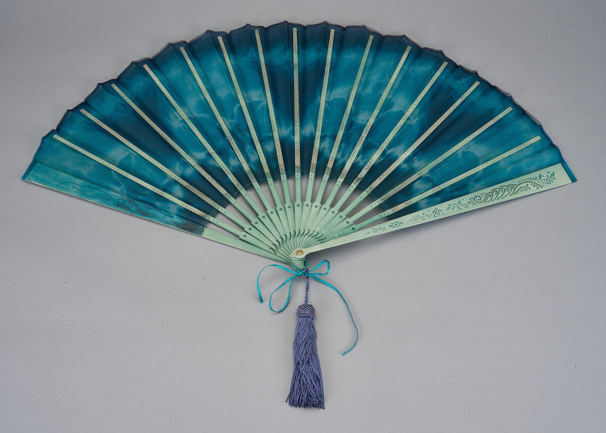 A decorative hand fan with blue and green hues, light wooden ribs, a blue tassel, and ribbon, displayed open against a plain gray background, evoking elegance and a tranquil, refined mood.