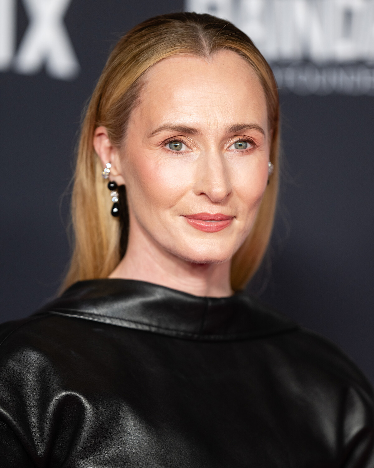 Woman with long blond hair wearing a black leather top and pearl earrings, standing in front of a dark blurred background at a formal event or premiere.