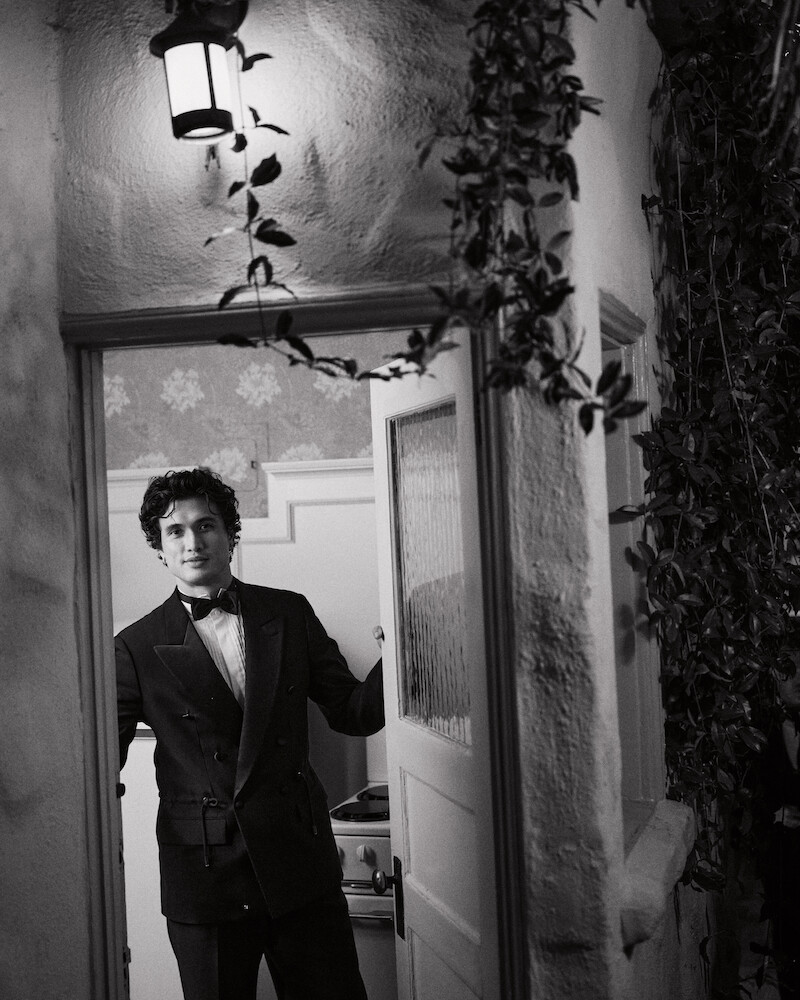 A man in a suit stands in a doorway, partially inside a house with patterned wallpaper, under a lit outdoor lantern, and surrounded by climbing plants in a black-and-white setting.