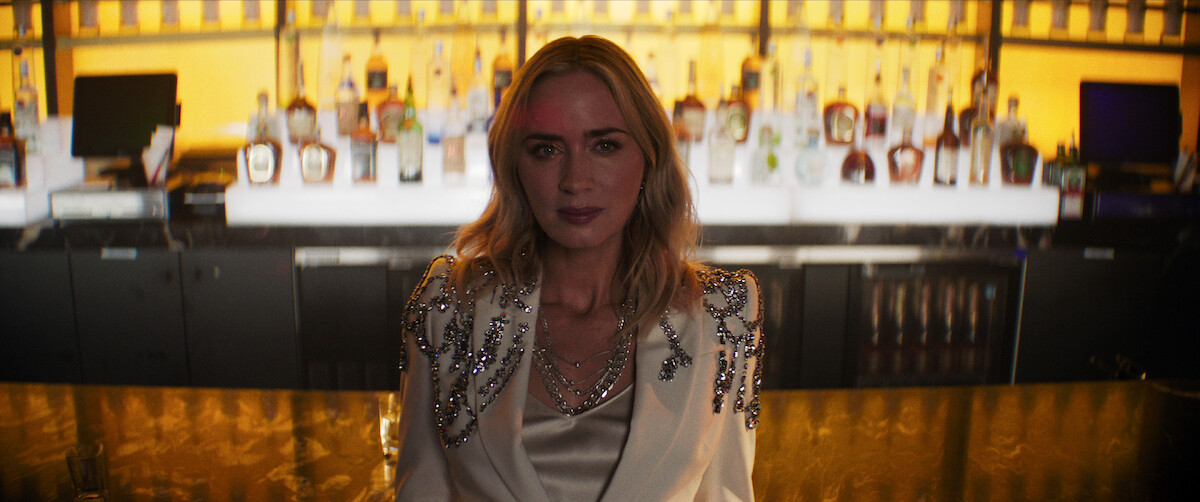 Emily Blunt as Liza sits in front of a bar.