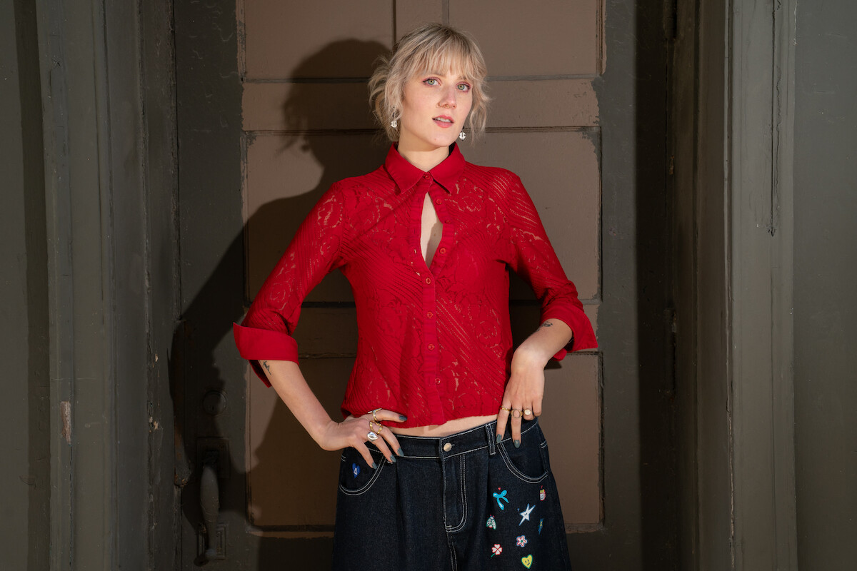 Person with short blonde hair wearing a red blouse and dark embroidered jeans, standing indoors in front of a brown door surrounded by gray painted walls with hands on hips, looking at the camera.