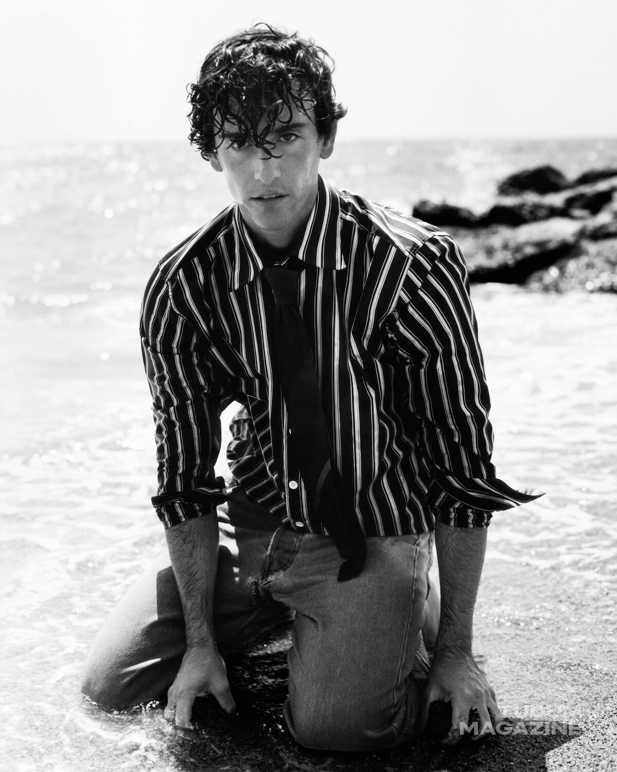 Man in striped shirt and tie kneeling on the wet sand at the beach, waves and rocks in the background, black and white photo, coastal environment, moody atmosphere.