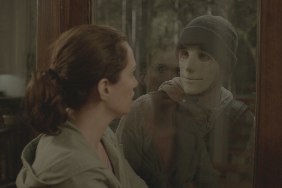 A woman inside a house faces a masked person outside looking in through a glass door, with the forest visible in the background, creating a tense and suspenseful atmosphere.