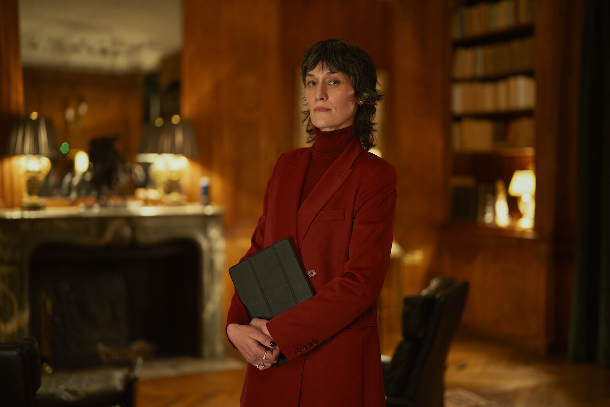 Uma mulher branca, alta, de cabelos curtos e pretos, veste um sobretudo vermelho em uma sala com iluminação quente e baixa