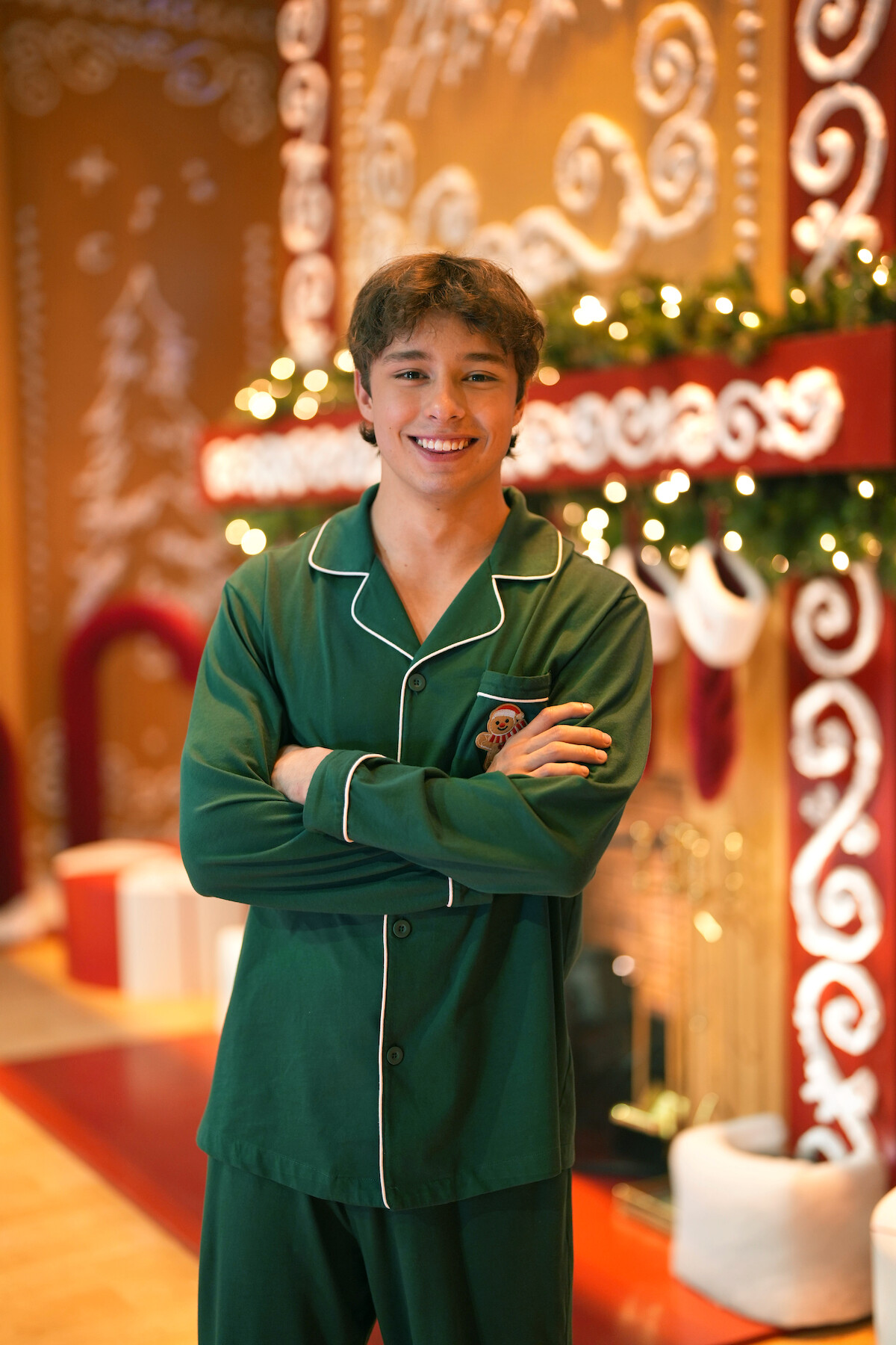 Smiling person in green pajamas stands with arms crossed in front of a festive holiday scene featuring gingerbread-style decorations, garlands, stockings, and gift boxes in a warmly lit room.