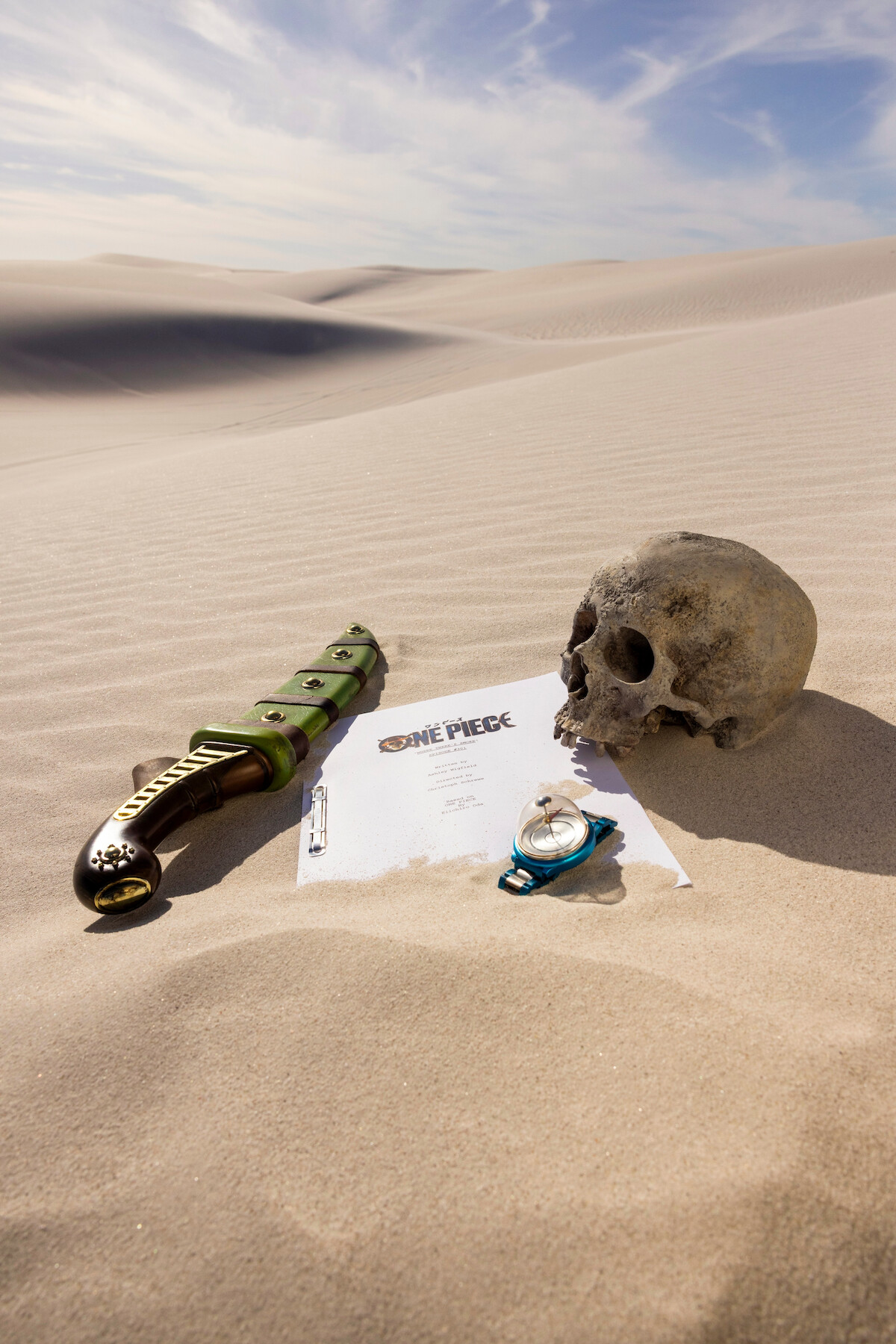 A skull, ornate sword, compass, and "One Piece" script page lie on sand dunes under a blue sky, suggesting an adventurous desert setting with themes of exploration and mystery.