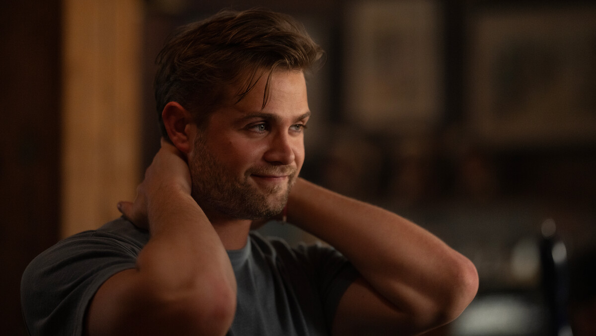 Leo Woodall as Vladimir with light brown hair smiling and resting hands behind his neck, sitting indoors with a relaxed expression, warm ambient lighting, and soft-focus framed pictures on a wall in the background.
