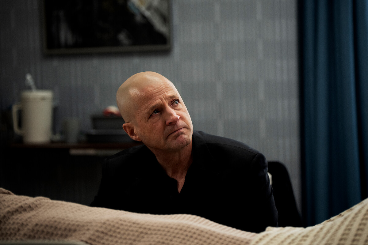 Bald man in a black suit sits beside a hospital bed in a dimly lit, somber room with medical equipment and a blanket in the foreground, looking up with a thoughtful, concerned expression.