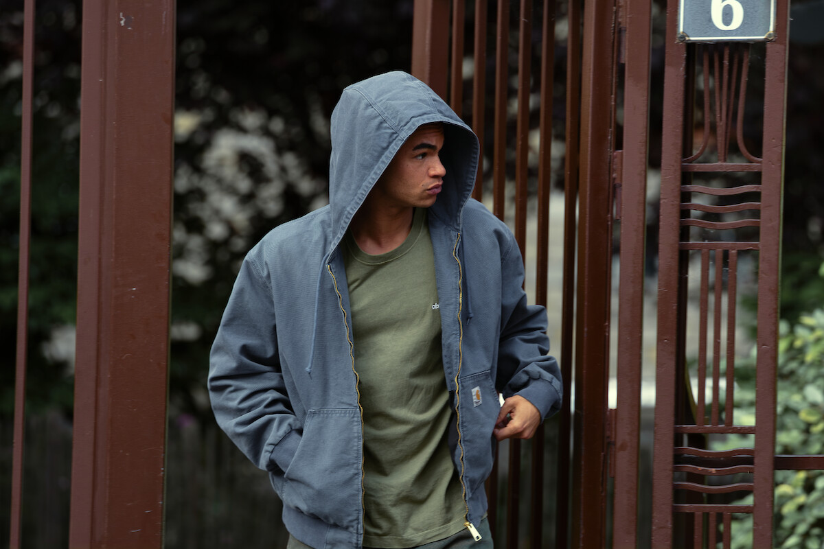 Um jovem negro usa camisa verde e um casaco azul com capuz cobrindo sua cabeça. Ele leva as mãos aos bolsos do agasalho e atravessa um portão de ferro marrom