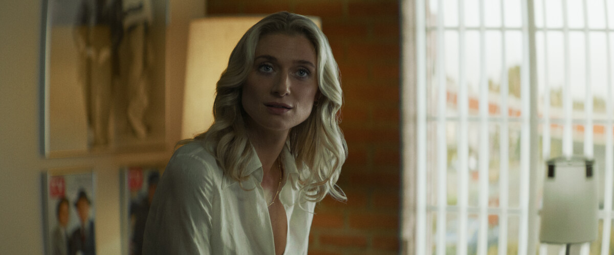 A woman with blonde hair sits indoors near a lamp and large window with vertical bars. The room has a brick wall, framed pictures, and soft lighting, giving a cozy, modern atmosphere.
