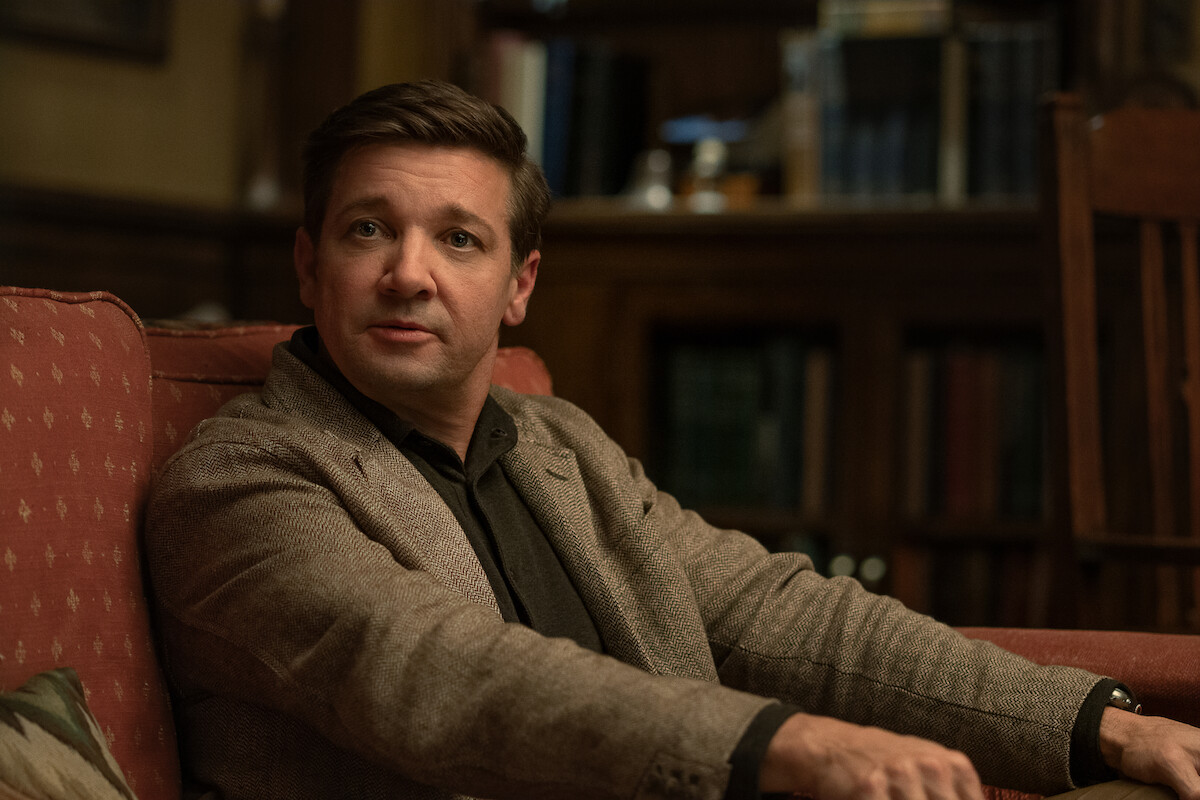 Man in a brown blazer sits on a red couch in a cozy, warmly lit library or study, surrounded by bookshelves, creating an atmosphere of calm conversation or thoughtful reflection.