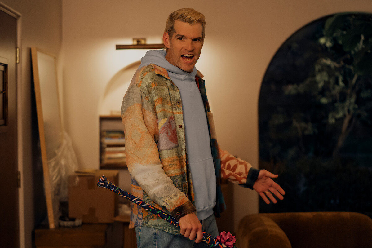 A man stands in a warmly lit living room holding a colorful umbrella with a pink handle, wearing a patterned jacket and light blue hoodie, near a brown couch and cardboard boxes, with a window showing trees outside at night.