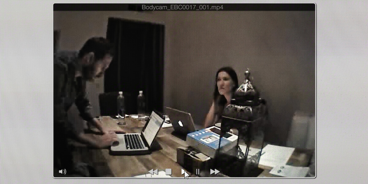 Two people working on laptops at a wooden table in a dimly lit room with water bottles, papers, and a decorative lantern. The scene appears on a computer screen as a paused video.