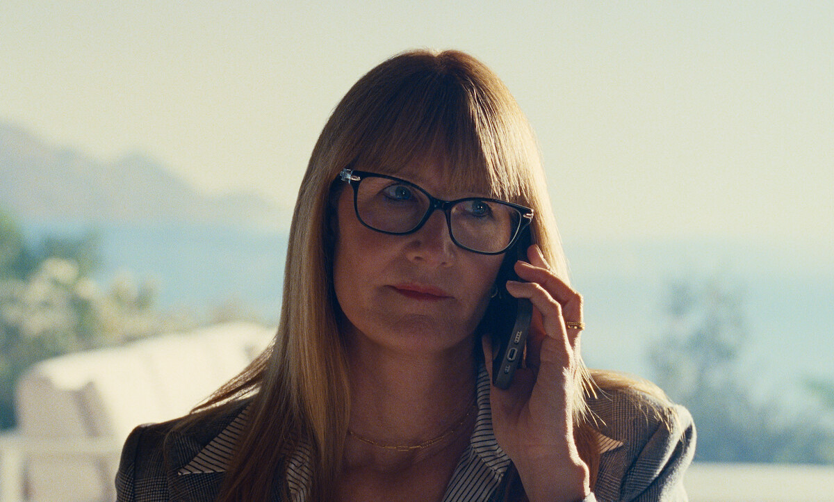 A woman with long hair and glasses talks on her cellphone in a softly lit, modern indoor setting, with blurred outdoor scenery and mountains visible in the background, creating a calm and focused mood.