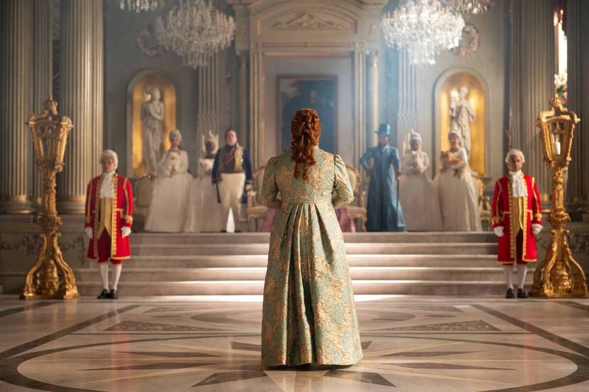A woman in an ornate gown stands in a grand, elegant hall facing a group in historical costumes on a staircase, surrounded by chandeliers and gold decor, evoking a royal or aristocratic setting.