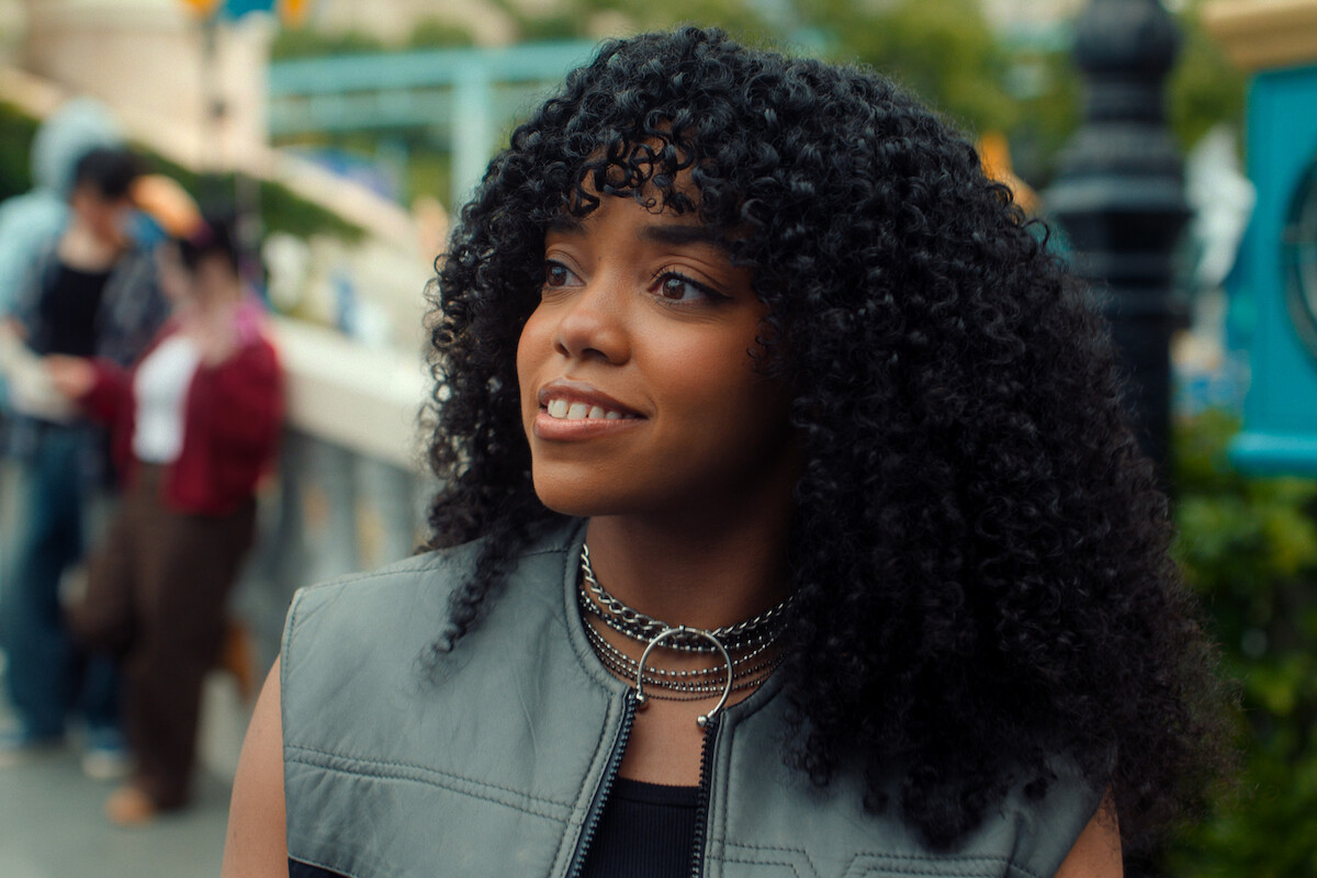 Young woman with curly hair smiling outdoors in an urban setting, wearing a gray vest and layered chain necklaces, with people and greenery blurred in the background.