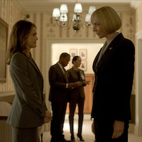 Two women in business suits face each other seriously in a formal, warmly lit office or hallway, while two people confer in the background under a chandelier, creating an atmosphere of tension and professionalism.