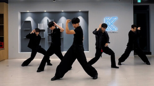 Five dancers in black outfits pose mid-routine in a modern dance studio with bright ceiling lights and a glowing K logo on the wall, light wooden floors, and geometric wall decor in the background.