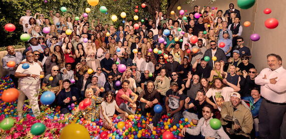 Large group of people of various ages gathered outdoors, posing in a ball pit with colorful plastic balls bouncing in the air; casual and joyful setting with trees and a wall in the background.