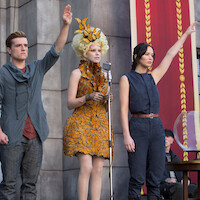 Three people stand on a stage in front of a microphone and banners, two with arms raised in a salute; urban setting, one in a decorative yellow dress, the others in simple outfits, evoking a ceremonial or public event atmosphere.