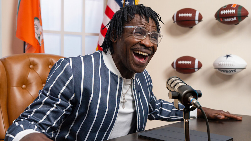 Man in striped jacket smiling and speaking into a microphone at a desk, with footballs mounted on the wall and flags in the background, resembling a sports interview or podcast setting.
