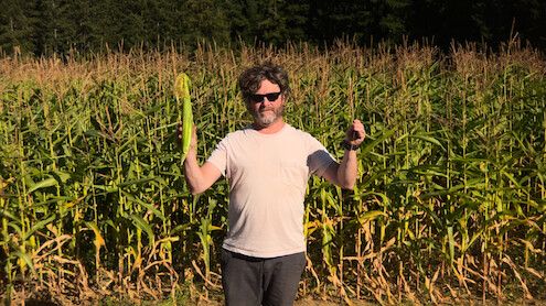 Zach Galifianakis stands in front of a corn field holding an ear of corn in one hand and a piece of wheat in the other.